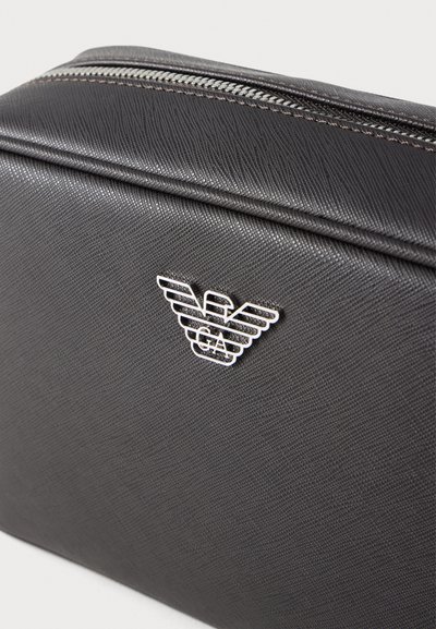 Μαύρη υφασμάτινη καλλυντική τσάντα με φερμουάρ και ασημένιο λογότυπο με φτερά. Το υλικό φαίνεται ανθεκτικό και λείο.