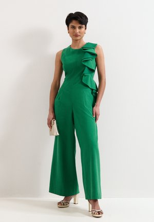 Frau trägt einen ärmellosen grünen Jumpsuit mit Rüschendetail auf einer Seite, hält eine beige Clutch und trägt beige Sandaletten mit Absatz.