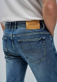 Jeans de denim azul con un acabado desgastado, que presentan una etiqueta de cuero etiquetada como "SALSA JEANS" en la cinturilla y bolsillos traseros con costuras sutiles.