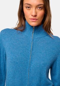 Pull zippé bleu clair en matériau doux, doté d'un col montant côtelé et de manches longues, avec une texture lisse sur l'ensemble.