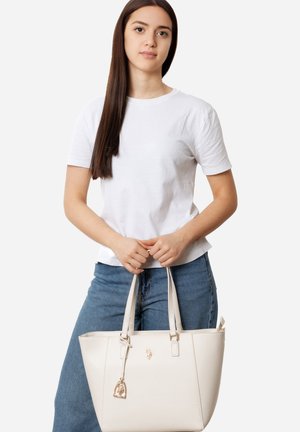 Joven con cabello largo y oscuro, vistiendo una camiseta blanca y jeans azules, sosteniendo un bolso tote grande color beige con asas dobles.