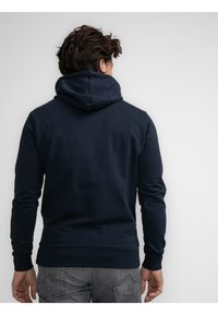 Marineblaue Kapuzenjacke mit großer Kapuze, langen Ärmeln und taillierter Passform. Hergestellt aus einem weichen, strukturierten Stoff. Die Rückansicht zeigt minimale Details.