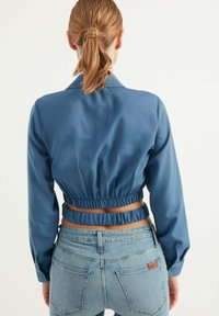 Camicetta blu corta con maniche lunghe, vita elastica e colletto. Indossata con jeans azzurri, che mostrano una texture liscia e un design aderente.