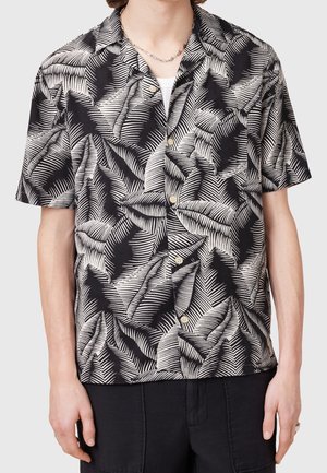 Mężczyzna noszący czarno-białą koszulę na krótki rękaw z abstrakcyjnym wzorem liści i srebrny naszyjnik z łańcuszkiem.