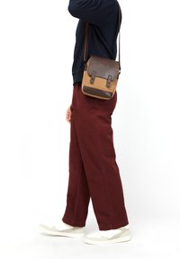 Bolso bandolera de cuero marrón y lienzo color canela con cierre de doble solapa, que presenta acentos metálicos, llevado sobre una blusa azul marino y pantalones burdeos.