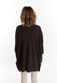 Lang, oversized donkerbruin sweater met een ronde halslijn, verlaagde schouders en een textuurstof. Gecombineerd met lichtgekleurde broeken.