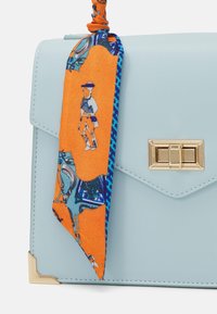Ljusblå läderhandväska med guldfärgad lås och hörnaccent, med en orange och blå mönstrad scarf knuten vid handtaget.