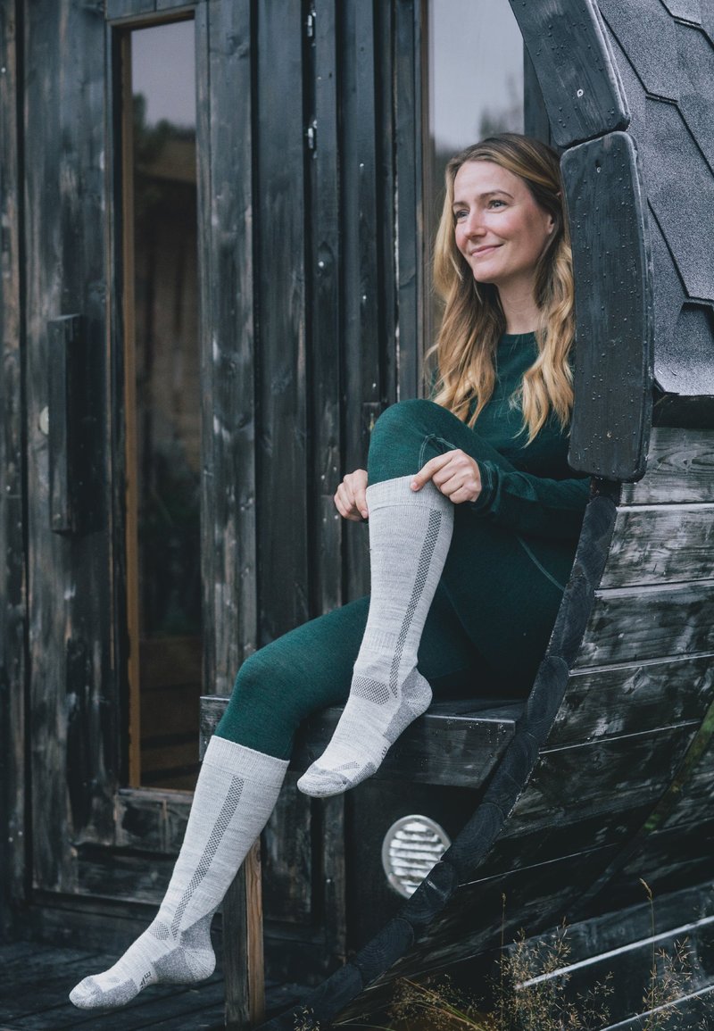 Grijze kniehoge sokken met een patroon, gedragen met een donkergroene outfit, zittend op een gebogen houten structuur. Natuurlijke omgeving op de achtergrond.