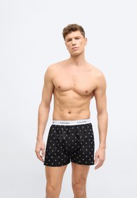 Άνδρας χωρίς πουκάμισο στέκεται μπροστά σε απλό φόντο φορώντας μαύρα μποξεράκια Calvin Klein με λευκή ζώνη μέσης με λογότυπο και μικρά λευκά σχέδια CK.