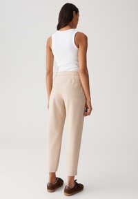 Femme portant un pantalon beige fuselé, un haut sans manches blanc et des baskets marron, debout de dos contre un fond clair.