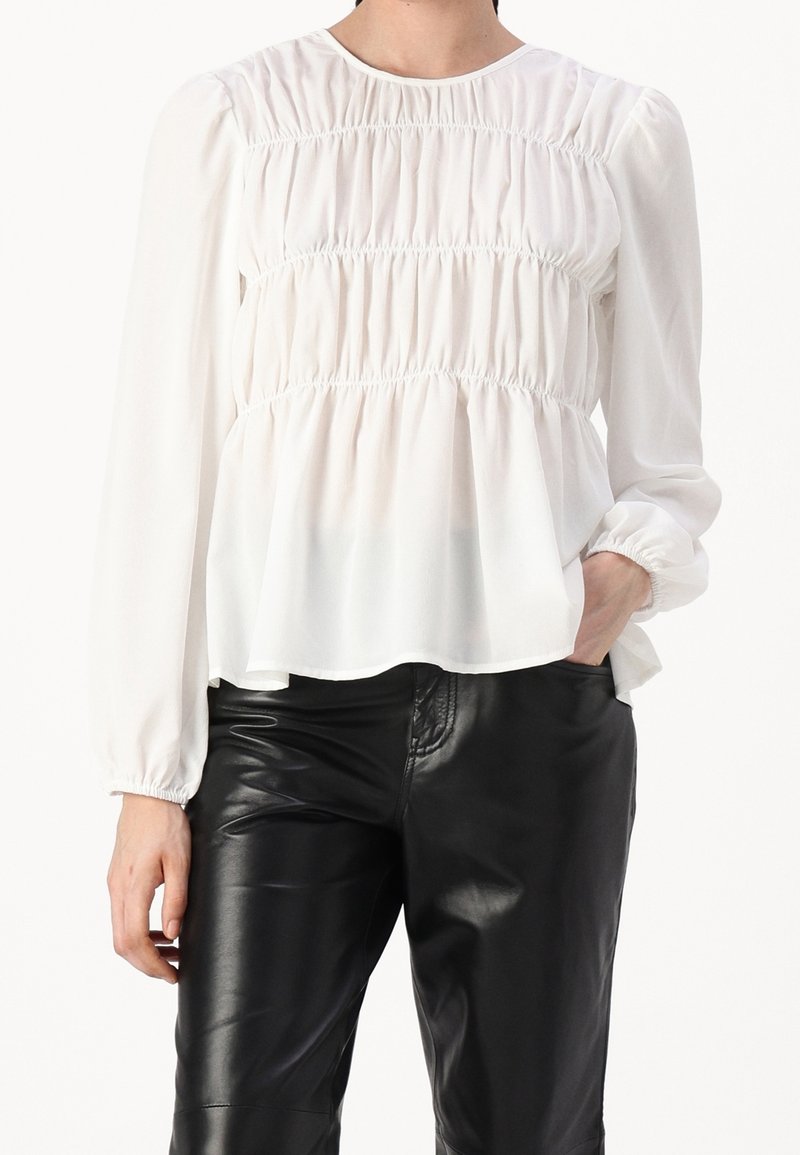 Blusa bianca a maniche lunghe con texture arricciata e scollo rotondo, abbinata a pantaloni di pelle nera. La blusa presenta una silhouette fluida.