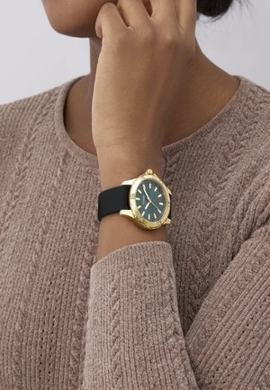 Gold wristwatch with a green dial and black silicone strap. Features include luminous hands and indexes, plus a textured bezel.