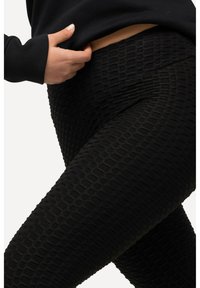 Personne portant des leggings noirs texturés et un haut noir à manches longues, main légèrement levée pour découvrir la taille.
