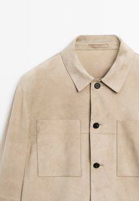 Beige suede jacket featuring a pointed collar, two chest pockets, and black buttons for closure. Smooth texture with a tailored design.