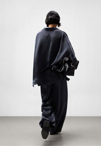 Cappa in satin blu navy con una vestibilità ampia, abbinata a pantaloni a gamba larga coordinati. La modella porta una borsa nera con texture, caratterizzata da un sottile dettaglio della tracolla.