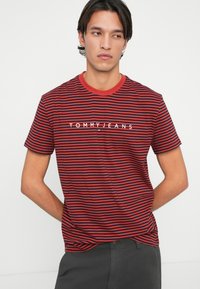 Rød og marineblå striped T-skjorte med rund hals, med en hvit "TOMMY JEANS" logo over brystet. Laget av et glatt, lett stoff.