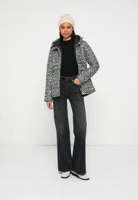 Zwart-wit gevlekte jas met capuchon, gecombineerd met een zwarte trui en hoog getailleerde uitlopende jeans. Roze gebreide muts en witte schoenen.