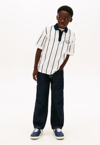 Λευκό πόλο πουκάμισο με ναυτικές ρίγες, κοντά μανίκια και γιακάς· συνδυασμένο με σκούρο μπλε παντελόνι ευρύχωλο και ναυτικά αθλητικά παπούτσια.