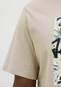 Camiseta de algodón beige con mangas cortas, que presenta un estampado floral y letras negras en negrita en el frente, textura suave y dobladillo recto.