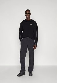 Camisola preta com um logótipo e costura branca contrastante; combinada com calças escuras e ténis desportivos pretos. Óculos de sol completam o visual.