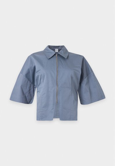 Veste courte bleu clair avec manches larges au niveau des coudes, fermeture éclair devant et col classique de chemise sur fond blanc.