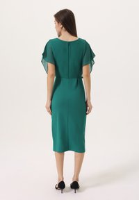 Robe midi verte avec des manches courtes en textile transparent, un col rond et une fermeture éclair dissimulée au dos. Texture lisse avec une silhouette ajustée.