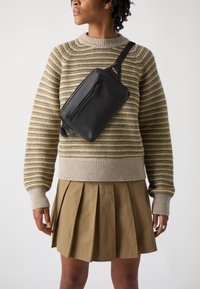 Sac en bandoulière en cuir noir avec une poche zippée à l'avant, porté en diagonale sur un pull en tricot rayé gris et jaune, associé à une jupe plissée beige.