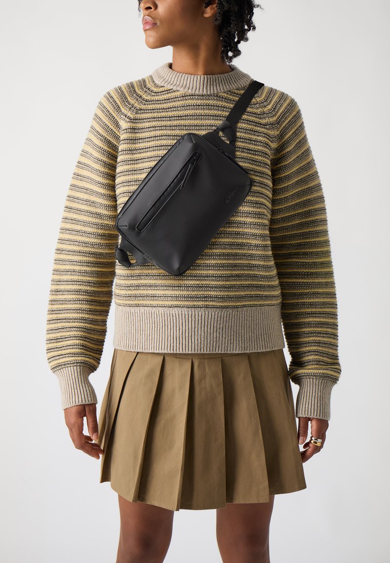 Sac en bandoulière en cuir noir avec une poche zippée à l'avant, porté en diagonale sur un pull en tricot rayé gris et jaune, associé à une jupe plissée beige.