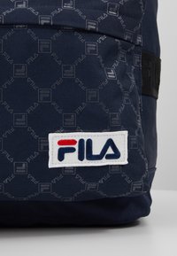 Námořnický modrý batoh s texturovanou tkaninou, který má opakovaný vzor loga FILA a bílou obdélníkovou nášivku s červeným a modrým nápisem.