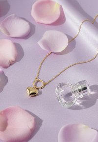 Collana con ciondolo a cuore d'oro e anello intrecciato, posata su raso lavanda con petali di rosa rosa, accanto a una bottiglia di profumo in vetro trasparente.