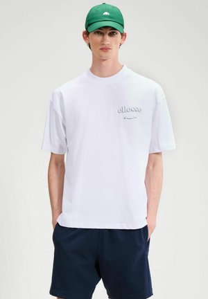 Mężczyzna w białej koszulce ellesse, granatowych szortach i zielonej czapce stoi z rękami w kieszeniach na jednolitym tle.