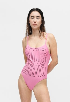 Žena s mokrými vlasmi v ružovom jednodielnom plavkách s veľkými nápismi "MOSCHINO", stojaca proti jednoliatemu svetlému pozadiu.