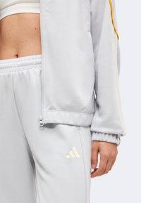 Κοντινό πλάνο ατόμου που φοράει λευκή φόρμα Adidas με το μπουφάν μερικώς φερμουαρ, εμφανίζοντας γυμνή κοιλιά και χέρι να ακουμπά στο πλάι.