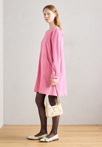 Ροζ ριγέ βαμβακερό φόρεμα με μακριά μανίκια και πλαϊνές τσέπες, συνδυασμένο με μαύρες καλσόν και κρέμα παπούτσια, κρατώντας μια κρέμα τσάντα με λεπτομέρεια φιόγκο.