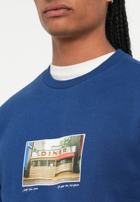 Close-up van een persoon in een blauwe sweatshirt met een afgedrukte foto van een vintage diner op de borst.
