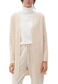 Kvinna som bär en beige öppen cardigan, vit polotröja och beige byxor med dragsko mot en vit bakgrund.