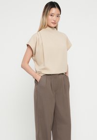 Top beige a maniche corte con colletto alto, abbinato a pantaloni marroni a vita alta con un design plissettato e tasche laterali.
