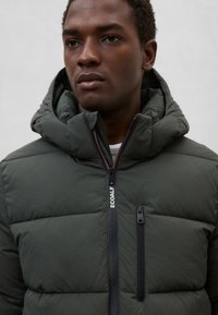 Mørkegrøn vatteret jakke med lynlås foran, høj krave og hætte. Har et logo på brystet og en lynlåslomme på venstre side.