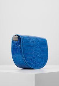 Borsa a tracolla in ecopelle blu con una finitura lucida a pelle di coccodrillo, forma curva e dettaglio in metallo sulla parte superiore.