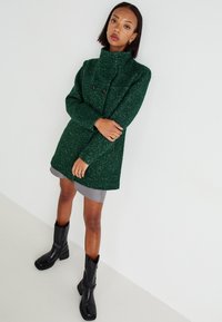 Manteau vert texturé avec col montant, double boutons noirs, et longues manches, associé à une jupe grise et des bottines noires.