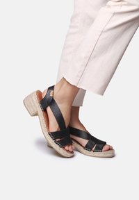 Crne sandale na remenčiće s potpeticom u stilu espadrille. Imaju tkanje od jute i glatku, teksturiranu gornju stranu.