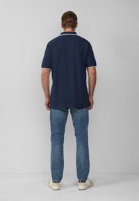 Man in een marineblauwe polo met witte accenten, blauwe jeans en witte sneakers, staand tegen een effen lichtgrijze muur.