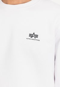 Hvit hettegenser med en liten svart Alpha Industries-logo på venstre brystområde mot en enkel bakgrunn.