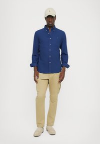 Jūras spēku pogām apģērbs, brūnu krāsu kravas bikses, bēša apavi un gaiši khaki cepure ar logo. Modelis pozē pret vienkāršu baltu fona.