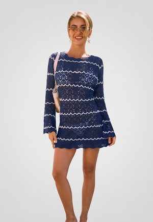 Femme souriante, portant une robe en dentelle bleue avec des rayures blanches ondulées, des lunettes de soleil, des boucles d’oreilles en forme d’étoile de mer et portant un sac à bandoulière beige.