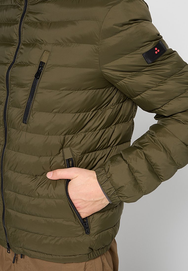 Olijfgroene pufferjack met een gewatteerd ontwerp, voorzien van ritszakken en een kleine zwarte patch op de schouder met rode accenten.