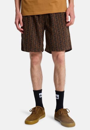 Braune Shorts mit Muster und vertikalen Designs, aus leichtem Stoff gefertigt. Getragen mit schwarzen Socken und braunen Wildlederschuhen.