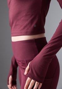 Maroon long-sleeve top with thumbholes and a logo, paired with high-waisted ribbed leggings. Smooth fabric with fitted design.