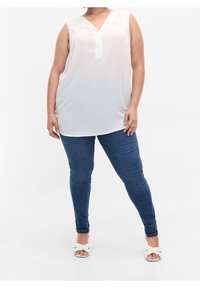 Blusa blanca sin mangas con escote en V y tejido semitranslúcido, combinada con jeans ajustados azul oscuro y sandalias de color claro.