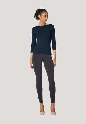 Kvinde med krøllet hår iført en navyblå top med trekvartærmer, sorte leggings og beige høje hæle, stående mod en ensfarvet lys baggrund.